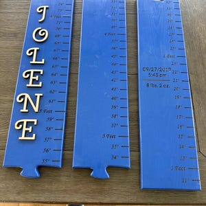 Puede incluir: Tres reglas de crecimiento de madera azul con medidas en pulgadas y pies. Una regla tiene el nombre "TOLENE" en letras doradas. Otra regla tiene la fecha, hora y peso de un bebé. Un artículo decorativo y personalizado.