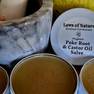 May include: Close-up of several small, round tins of organic salve, labeled "Poke Root & Castor Oil Salve" with a shamrock symbol. A marble mortar and pestle are in the background, along with dried herbs.