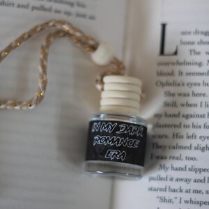 May include: A small glass bottle with a black label that reads "In My Dark Romance Era". The bottle is hanging from a white and gold cord and is on top of a book.
