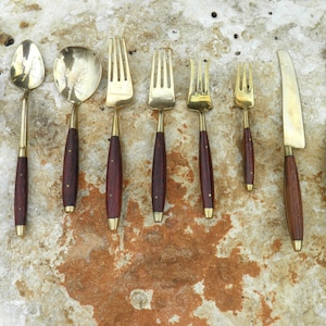 May include: A set of brass and wood cutlery, including a spoon, fork, and knife. The cutlery has a dark brown wood handle and a gold-colored metal head.