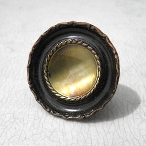 May include: A decorative, antique-style knob with a black outer rim and a brass-colored center. The center is surrounded by a twisted rope design. The knob has a scalloped edge and is likely for a drawer or cabinet.