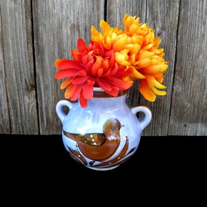 May include: A small, light blue ceramic vase with a brown bird design and two handles. The vase holds vibrant orange and red artificial flowers. The background is a weathered wooden surface, and the vase sits on a black surface.