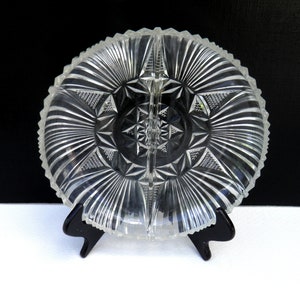 May include: Clear, divided glass serving dish with a starburst and geometric design. The dish has a scalloped edge and is supported by a black stand. The dish is divided into two sections, perfect for serving snacks or appetizers.