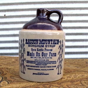 May include: A brown and beige ceramic jug with a blue label that reads "Racoon Mountain Sorghum Syrup Open Kettle Process Made On Our Farm Net. Wt. 1 Qt., 32 Fluid Oz. Delicious on Pancakes-Waffles-Biscuits-Cornbread Fritters-Hot French Bread-Hot Rolls French Toast Old Fashioned Made From Cane Renheth Farmer, Pisgah, Alabama".