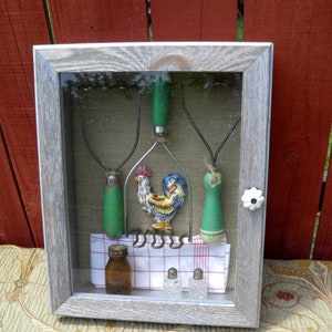May include: A rustic wooden shadow box with a whitewashed finish, displaying a collection of vintage kitchen tools. The box features three green potato mashers, a rooster-shaped metal utensil, and two small glass salt shakers. A red and white checkered cloth is placed at the bottom of the box.