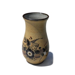 May include: A small, tan ceramic vase with a brown rim. The vase is decorated with a black and brown bird and floral design.