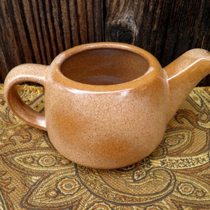 May include: A tan ceramic teapot with a handle and spout. The teapot has a speckled texture and sits on a gold paisley patterned fabric. The background is a dark wooden surface.