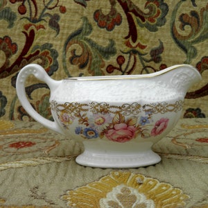 May include: A white ceramic gravy boat with a gold trim and floral design. The boat has a handle and a spout.