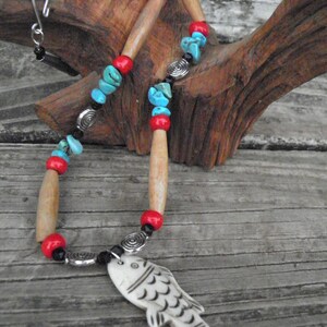 May include: A handcrafted necklace featuring a carved white fish pendant. The necklace incorporates wooden beads, turquoise chips, red beads, and silver-toned spiral accents. The necklace is displayed on a wooden surface.