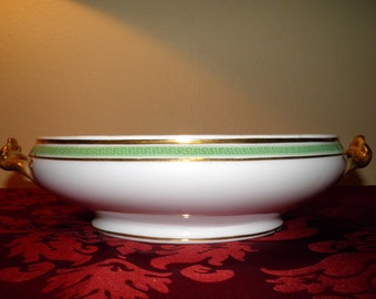 May include: A white porcelain serving bowl featuring a gold rim and a decorative green Greek key pattern band. The bowl has two gold-toned handles on opposite sides and sits on a circular pedestal base with a thin gold line accenting the bottom edge.