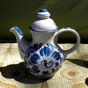 May include: A white ceramic teapot with a blue floral design. The teapot has a curved spout, a rounded handle, and a lid with a blue finial. The floral pattern is painted in shades of blue and white.