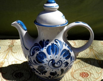 May include: A white ceramic teapot with a blue floral design. The teapot has a curved spout, a rounded handle, and a lid with a blue finial. The floral pattern is painted in shades of blue and white.