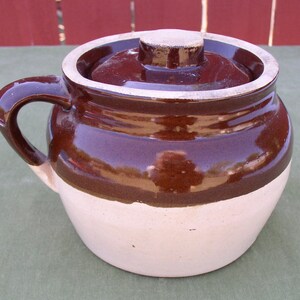 May include: A brown and white glazed ceramic crock with a lid. The crock has a rounded body and a single handle.