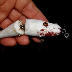 May include: A vintage white and red fishing lure with a segmented body and metal hardware. The lure has a weathered appearance with red paint and a hook. The lure is held in a hand against a black background.