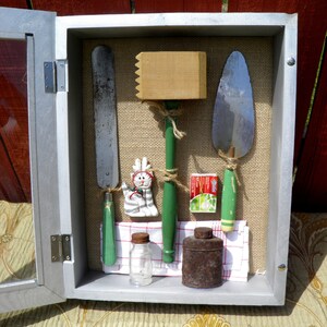 May include: A vintage wooden box with a glass door, containing a collection of tools and objects. The box is painted silver and has a burlap background. The tools include a wooden mallet, a metal spatula, a metal scraper, and a small glass bottle. There is also a small ceramic figurine of a cat and a small metal container.