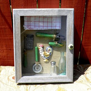 May include: A gray wooden shadow box with a glass front, displaying a collection of vintage kitchen tools and a rooster figurine. The tools include a rolling pin, a grater, a pizza cutter, and a bottle opener. The rooster is painted in bright colors and has a red comb and wattle.