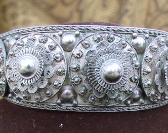May include: A silver-toned metal bracelet with intricate floral and circular designs. The bracelet features a series of large, domed centerpieces surrounded by smaller, detailed floral patterns. The edges are adorned with a braided rope-like texture.