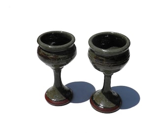 May include: Two handmade ceramic wine goblets. The goblets are dark green with brown and red accents. The stem and base are a lighter green, and the base has a red band. The interior of the goblets is dark.