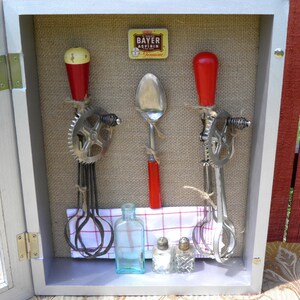 May include: A white wooden shadow box with a burlap background. Inside the box are three vintage kitchen tools with red handles, a small bottle, and two salt shakers. The tools are a spoon, a hand-crank egg beater, and a hand-crank ice cream scoop. The box also contains a small yellow box with the text "Bayer Aspirin Genuine".