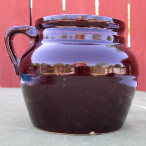 May include: A dark brown glazed pottery crock with a single handle. The crock has a rounded body and a slightly flared rim.