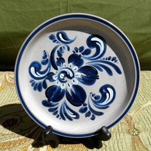 May include: A round ceramic plate with a floral design. The plate has a light gray center with a dark blue border and a dark blue and white floral pattern in the center. The plate is displayed on a black stand.