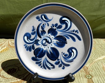 May include: A round ceramic plate with a floral design. The plate has a light gray center with a dark blue border and a dark blue and white floral pattern in the center. The plate is displayed on a black stand.