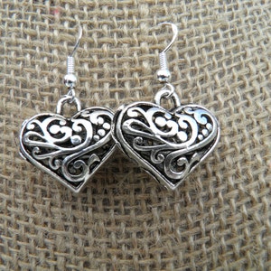 May include: A pair of silver-colored heart-shaped earrings. Each earring features an openwork design with intricate swirls and patterns. The earrings hang from a simple hook.