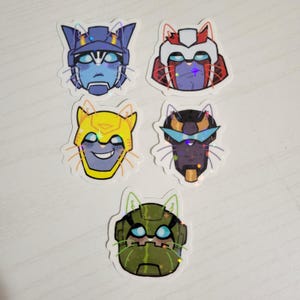 May include: A collection of five stickers featuring robot-cat hybrids. Each sticker has a different color scheme, including blue, red, yellow, black, and green. The stickers have a holographic, glittery effect.