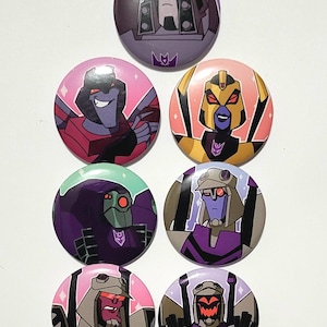Op de afbeelding: Een verzameling van zeven ronde buttons met cartoon illustraties van Transformers karakters. De buttons tonen verschillende karakters in levendige kleuren, waaronder paars, roze, goud en groen, met het Transformers logo.