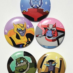 Op de afbeelding: Vijf ronde buttons met cartoonkarakters uit de Transformers-franchise. De buttons hebben verschillende kleuren, waaronder blauw, geel, roze, groen en goud. Elke button toont het gezicht van een ander personage.