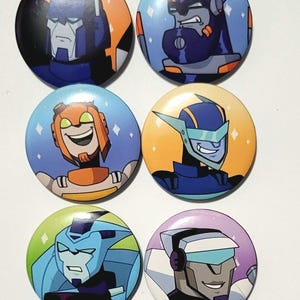 Op de afbeelding: Zes ronde buttons met cartoon robotkarakters in verschillende kleuren. De buttons hebben blauwe, oranje, groene en paarse achtergronden. Elke button toont een ander robotkarakter met unieke gezichtsuitdrukkingen en ontwerpen.