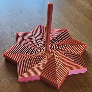 Peut inclure: Objet en forme d'étoile imprimé en 3D, dans des tons de rose et de rouge. La base présente un design en couches, avec une tige verticale centrale. L'objet est en plastique et repose sur une surface en bois.