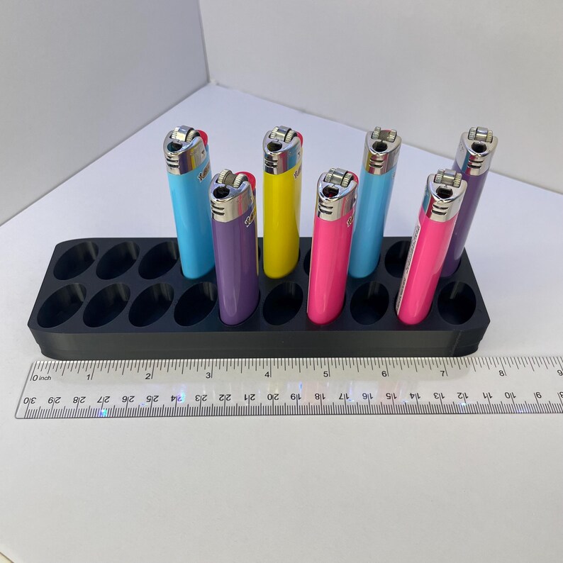 May include: A black plastic organizer with twelve oval slots holding six colorful lighters in blue, purple, yellow, pink, and violet. A clear plastic ruler is positioned in front, showing the organizer is approximately 9 inches in length.