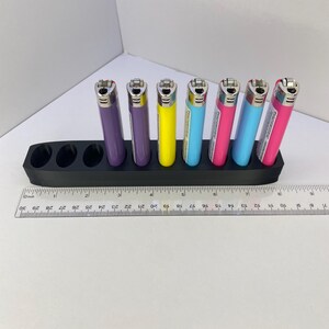 Pu&ograve; includere: Un supporto in plastica nera con nove fori circolari, contenente sei accendini colorati in viola, giallo, azzurro e rosa. Un righello in plastica trasparente &egrave; posizionato davanti, indicando che il supporto &egrave; lungo circa 19 cm.