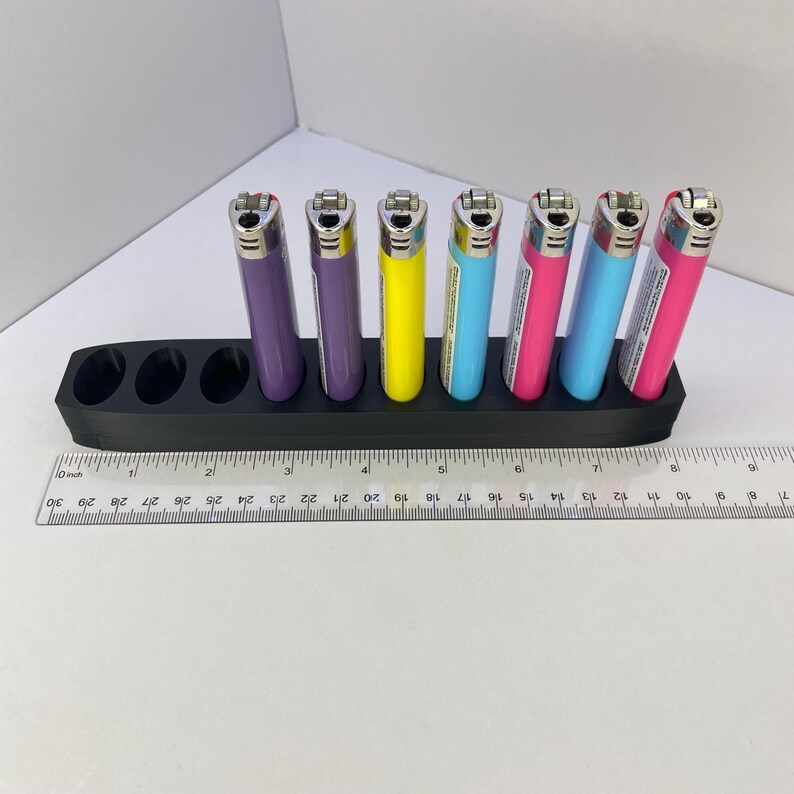 Pu&ograve; includere: Un supporto in plastica nera con nove fori circolari, contenente sei accendini colorati in viola, giallo, azzurro e rosa. Un righello in plastica trasparente &egrave; posizionato davanti, indicando che il supporto &egrave; lungo circa 19 cm.