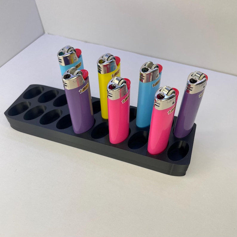May include: A black plastic display tray with multiple oval slots holding six colorful Bic lighters. The lighters are in shades of purple, yellow, pink, and light blue, each featuring a metallic top with a spark wheel and the Bic logo printed on the side.