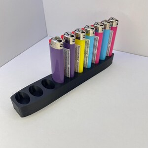 Pu&ograve; includere: Un supporto organizzatore rettangolare in plastica nera con otto fori circolari, contenente sette accendini usa e getta colorati nelle tonalit&agrave; viola, giallo, azzurro e rosa. Il supporto presenta tre fori vuoti nella parte anteriore.
