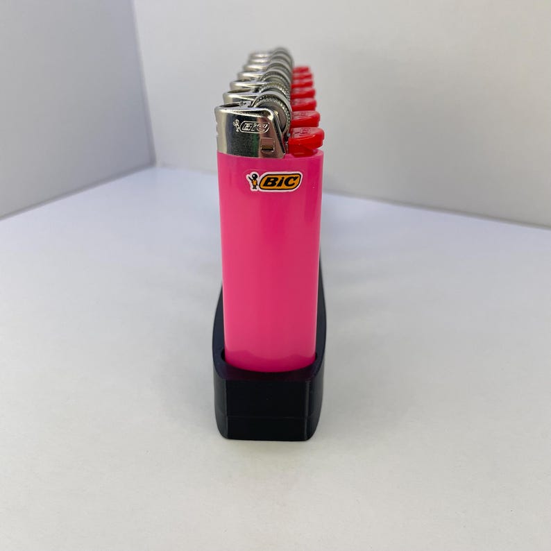Pu&ograve; includere: Una fila di accendini BIC rosa brillante con cime in argento metallizzato e pulsanti di accensione rossi, tenuti in posizione verticale in un supporto rettangolare nero. Ogni accendino presenta il logo BIC giallo e nero sul corpo rosa.