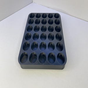 Pode incluir: Uma bandeja de armazenamento retangular preta com uma grade de trinta aberturas circulares dispostas em cinco linhas e seis colunas. A bandeja tem um acabamento fosco liso e paredes verticais uniformes para pequenos objetos.