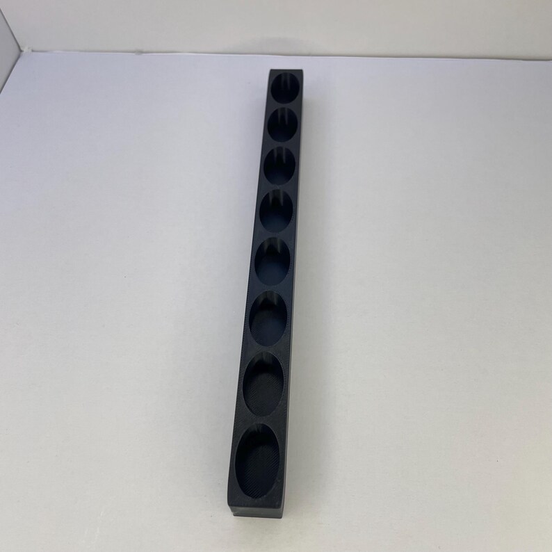 Op de afbeelding: Een lange, smalle, matzwarte rechthoekige organizer-tray met negen gelijkmatig verdeelde, ronde uitsparingen in een enkele verticale rij. Het object heeft een strak, gestructureerd ontwerp met gladde randen en een uniforme donkere afwerking.