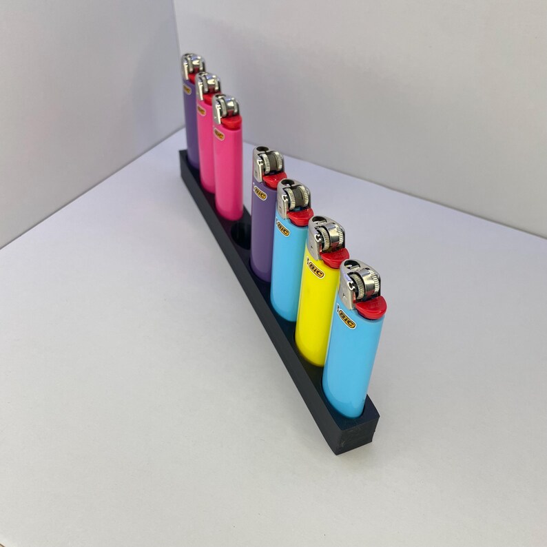Op de afbeelding: Een zwarte rechthoekige standaard met zeven Bic-aanstekers op een rij. De aanstekers hebben verschillende effen kleuren, waaronder paars, roze, lichtblauw en geel, elk met een metalen ontstekingsmechanisme en een rode knop met het Bic-logo.