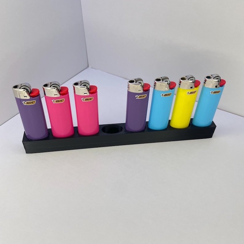 Op de afbeelding: Een zwarte, rechthoekige kunststof houder met zes Bic-aanstekers in diverse kleuren, waaronder paars, roze, lichtblauw en geel. De aanstekers staan in individuele sleuven, met &eacute;&eacute;n lege ronde opening zichtbaar in het midden van de basis.