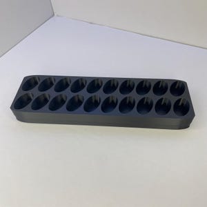 May include: A rectangular dark gray storage tray featuring two parallel rows of twelve oval-shaped recessed compartments. The tray has rounded corners and a smooth, matte finish, designed to hold small items in its twenty-four individual slots.