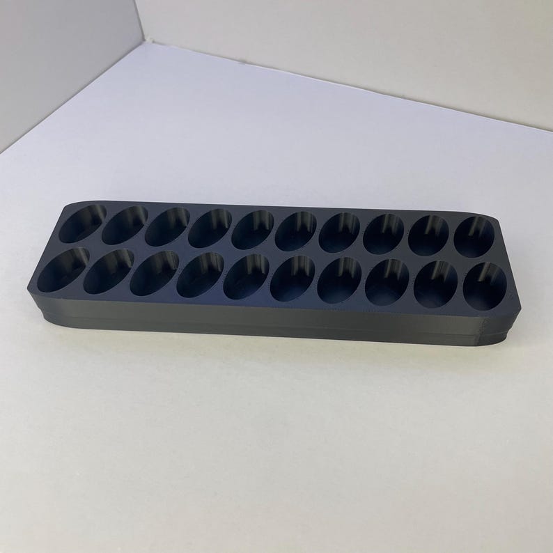 May include: A rectangular dark gray storage tray featuring two parallel rows of twelve oval-shaped recessed compartments. The tray has rounded corners and a smooth, matte finish, designed to hold small items in its twenty-four individual slots.