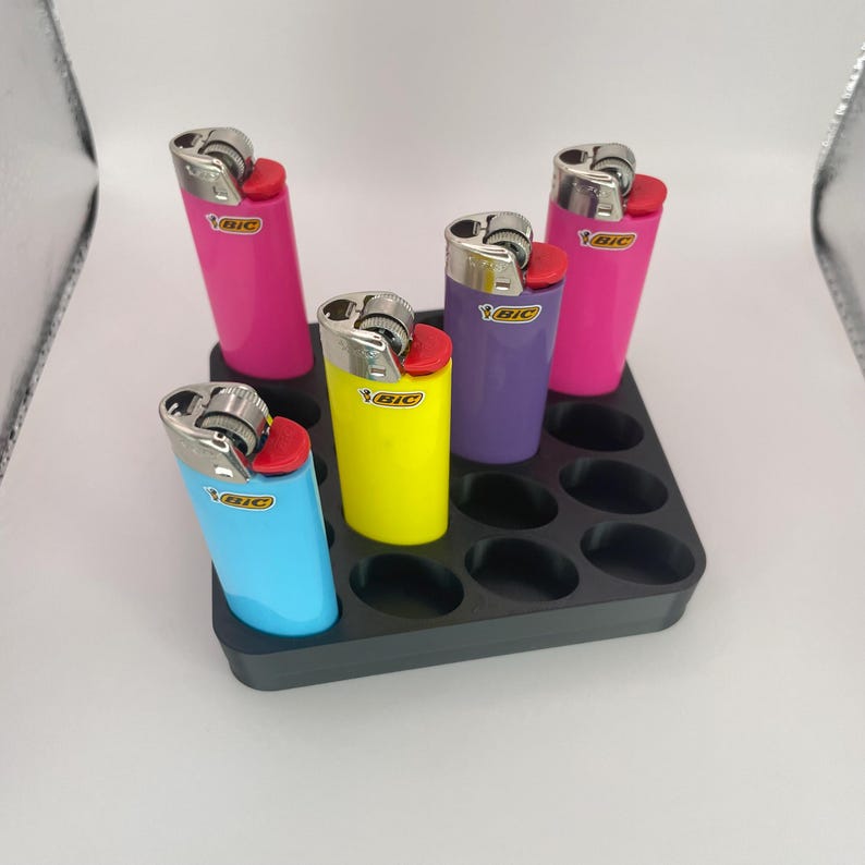 May include: A black tray holds five colorful lighters: pink, yellow, purple, and blue. Each lighter has a silver metal top and a red ignition button. The lighters are arranged in a grid pattern.
