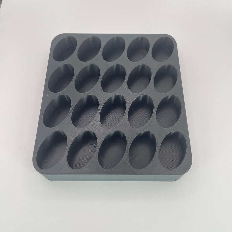 May include: A dark gray, rectangular organizer with 24 oval-shaped compartments. The organizer is made of a solid material and is designed to hold small items. The compartments are evenly spaced and have rounded edges.