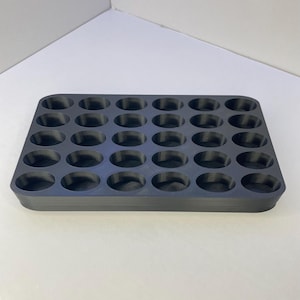 Pode incluir: Uma bandeja de armazenamento retangular preta com uma grade de trinta compartimentos circulares embutidos, dispostos em cinco fileiras de seis. A bandeja tem cantos arredondados e um acabamento fosco liso, projetado para guardar pequenos itens.