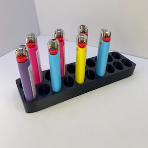May include: A black rectangular plastic organizer stand with multiple circular slots, holding five colorful Bic lighters in purple, pink, light blue, yellow, and blue. The lighters feature metallic ignition wheels and red buttons.