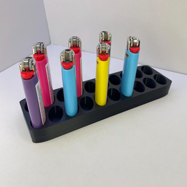 May include: A black rectangular plastic organizer stand with multiple circular slots, holding five colorful Bic lighters in purple, pink, light blue, yellow, and blue. The lighters feature metallic ignition wheels and red buttons.