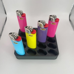May include: A black plastic organizer holding five colorful BIC lighters. The lighters are in various colors: pink, blue, yellow, and purple. Each lighter has a red ignition button and a silver metal top. The organizer has 12 oval-shaped slots.